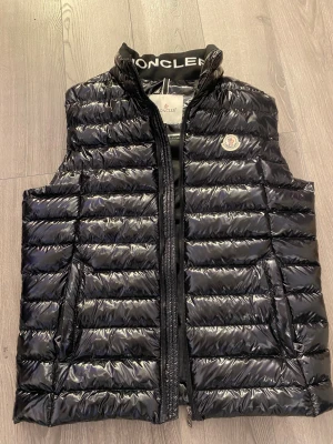 Svart pufferväst från Moncler (Storlek M) - Snygg svart pufferväst från Moncler med glansig finish och broderad logga på bröstet. Västen har ståkrage med Moncler-tryck, horisontella quiltade paneler och dragkedja framtill. Perfekt för lager-på-lager och streetstyle. AA+