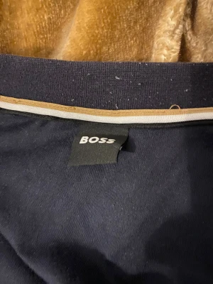 Svart bomberjacka från BOSS - Snygg svart bomberjacka från BOSS med gråa detaljer och dragkedja framtill. Jackan har ribbade muddar med vita och beige ränder vid hals, ärmslut och nederkant. Perfekt för dig som gillar stilren och sportig look.