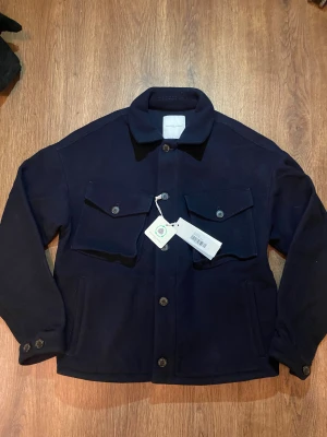 Mörkblå overshirt från Philippe Model - Stilren mörkblå overshirt från Philippe Model Paris. Jackan har klassisk krage, två stora bröstfickor med lock och knappar samt raka ärmar. Tillverkad i mjuk ullmix som ger en exklusiv känsla. Perfekt för lager-på-lager och snygg till jeans eller chinos.