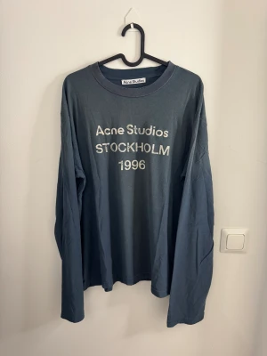 Blå långärmad topp Acne Studios storlek M - Säljer en blå långärmad topp från Acne Studios med tryck 'Acne Studios STOCKHOLM 1996' på bröstet. T-shirten har rund halsringning och är tillverkad i mjuk bomull. Perfekt för en avslappnad och cool look.