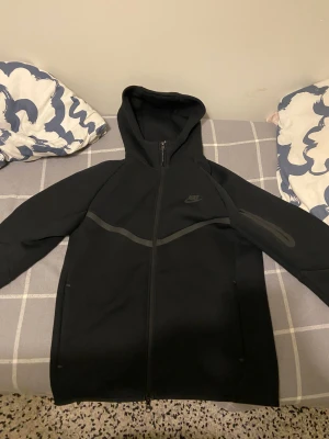 Nike tech fleece - Svart Nike tech med stilren design och huva. Jackan har hel dragkedja framtill, diskret Nike-logga på bröstet och en snygg mörkgrå detalj över bröstet. Ärmarna är långärmade och har en extra ficka med dragkedja på ena armen. Perfekt för dig som gillar sportig stil.
