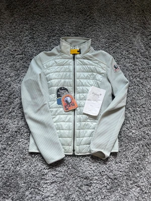 Ljusgrå quiltad jacka Parajumpers S - Snygg ljusgrå quiltad jacka från Parajumpers med dragkedja och hög krage. Jackan har ett sportigt uttryck, coolt gult spänne vid halsen och klassisk PJS-logga på ärmen. Materialmix med glansig front och strukturerade ärmar. Perfekt för lager-på-lager under kyliga dagar.
