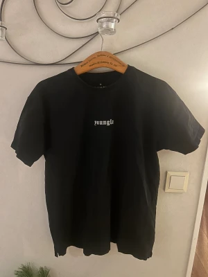 Svart t-shirt från YoungLA - Svart t-shirt från YoungLA med vit text på bröstet. Klassisk rund halsringning och korta ärmar. Tillverkad i mjuk bomull för en skön känsla och avslappnad passform. Perfekt för en stilren och enkel look.