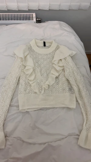 Vit stickad tröja med volanger - Supersöt vit stickad tröja från H&M med snygga volangdetaljer på axlar och framstycke. Tröjan har ett stickat genombrutet mönster. Oanvänd!