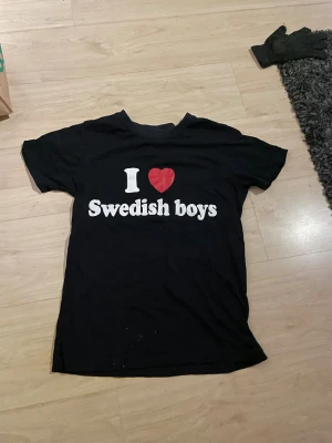 Svart t-shirt med I ❤️ Swedish boys-tryck - Svart t-shirt med vit och röd text på bröstet där det står 'I ❤️ Swedish boys'. Klassisk passform och rund halsringning. Perfekt statement-plagg för dig som vill sticka ut och visa din kärlek till svenska killar. storlek MAN S