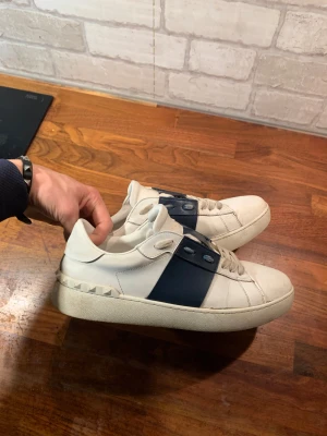 Valentino Garavani Open sneakers vit/blå - Snygga Valentino Garavani Open sneakers i vitt skinn med bred marinblå läderdetalj över snörningen. Klassisk rund tå, platt sula och ikoniska nitar längs hälen. Perfekta för dig som vill ha en stilren men ändå lyxig look.