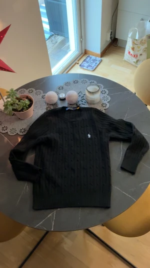 Svart kabelstickad tröja Polo Ralph Lauren - Svart kabelstickad tröja från Polo Ralph Lauren i storlek S. Klassisk design med rund hals och broderad vit logga på bröstet. Tröjan har långa ärmar och ribbade muddar vid ärmslut och nederkant. Perfekt för dig som gillar stilrena och tidlösa plagg.