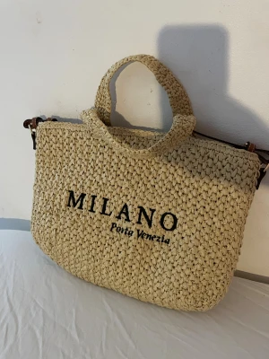 Beige handväska Milano H&M - Snygg beige handväska från H&M med texten 'MILANO Porta Venezia' broderad på framsidan. Väskan är flätad i ett naturmaterial och har både handtag och axelrem i brunt. Perfekt för dig som vill ha en trendig och rymlig väska med sommarkänsla.