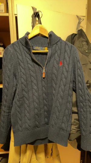 ralph lauren quater zip - Snygg mörkblå stickad tröja från Polo Ralph Lauren med klassiskt flätmönster och halv dragkedja framtill. Tröjan har ribbad krage, muddar och det ikoniska röda Polo-logot på bröstet. Perfekt för dig som gillar stilren och tidlös design.