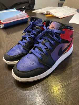 Nike Air Jordan 1 Mid City Sunset - Nike Air Jordan 1 Mid med coolt city sunset-motiv på sidan. Färgerna är lila, blå, svart och röd med matchande blå snören. Skorna har en klassisk rund tå, snörning och vit platt sula med transparent orange-blå undersida. Materialet är skinn och syntet. Använda 1-2 gånger Låda och allt man fick med fins kvar och är hela.