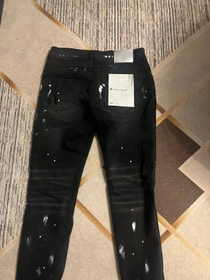 Svarta skinny jeans från Purple Brand - Svarta jeans från Purple Brand med skinny passform och coola vita färgstänk samt slitna detaljer vid knäna. Jeansen har klassisk femficksdesign, knappgylf och etikett bak. Materialet är stretchigt jeans för skön känsla och snygg look.