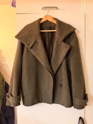 Olivgrön kappa från ARKET - Säljer en olivgrön kappa från ARKET med bred krage och dubbelknäppning. Jackan är tillverkad i ull från Manteco, har långa ärmar med knappdetalj vid mudden och en loose passform. Använd ett antal gånger, men får inte längre användning av den💗