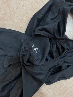 Svart baddräkt från SOC, storlek 38 - Sportig svart baddräkt från SOC med snyggt brottarrygg och diskret rynkdetalj vid bysten. Materialet är stretchigt syntet som sitter bekvämt och torkar snabbt. SOC-logga i vitt på höften ger en clean look. Perfekt för dig som gillar att simma eller hänga vid poolen.