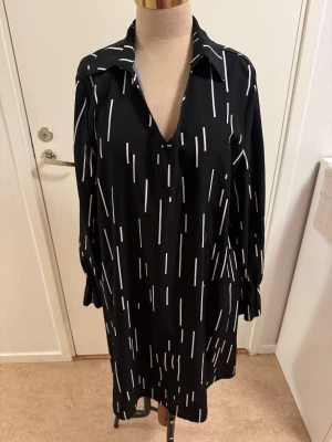 Svart-vit mönstrad skjortklänning SHEIN - Svart skjortklänning från SHEIN i storlek L med vita vertikala streck som mönster. Klänningen har lång ärm, klassisk krage och v-ringning framtill. Den har en loose passform och är tillverkad i ett mjukt, lätt material som känns bekvämt mot huden.