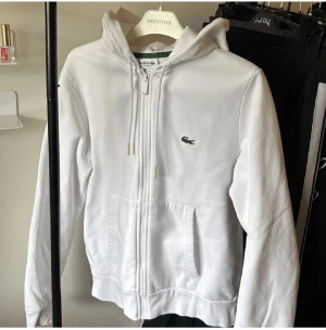 Vit hoodie från Lacoste med dragkedja - Vit hoodie från Lacoste med klassisk krokodillogga på bröstet. Tröjan har huva med snörning, hel dragkedja framtill och två fickor. Perfekt för en chill och sportig stil.