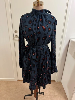 Mörkblå leopardklänning från SHEIN - Mörkblå kortklänning från SHEIN med leopardmönster i svart och rött. Klänningen har lång ärm, hög hals med knytning i nacken, markerad midja med bälte och volangdetaljer i kjolen. Tillverkad i ett lätt och skönt polyestertyg.