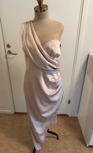 Champagnefärgad one shoulder-klänning - Säljer en elegant champagnefärgad långklänning från Lavish Alice. Klänningen har en draperad design med one shoulder och är tillverkad i glansigt satinliknande material. Dold dragkedja bak och snyggt omlottfall framtill för en lyxig look.