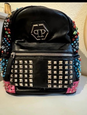 Philipp Plein svart ryggsäck med nitar - Cool svart ryggsäck från Philipp Plein i skinn med massor av färgglada nitar och silverfärgade detaljer. Väskan har en stor ficka framtill med dragkedja, quiltad baksida och rosa metallic handtag. Logotypen i silver på framsidan ger extra edge.lädret är dåligt men väskan är inte trasigt. Priset kan diskuteras ☺️