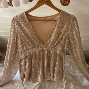 Champagnefärgad paljettopp från Nelly  - Glittrig champagnefärgad topp från NELLY med v-ringning och långa ärmar. Toppens yta är täckt av paljetter som ger ett riktigt festligt intryck. Modellen är lite lösare och har ett markerat band under bysten som man kan knyta för en snygg siluett.(säljer denna på vinted oxå💕) (köpte den för 300/400,bara använd en gång där av priset)🥹