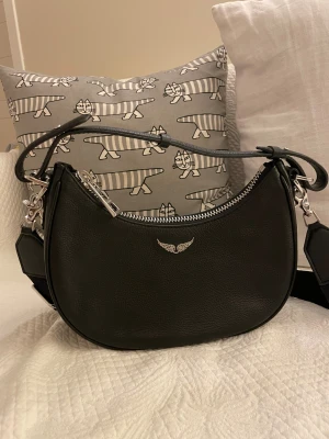 Svart axelväska från Zadig & Voltaire - Snygg svart axelväska från Zadig & Voltaire i äkta skinn med silverfärgad dragkedja och ikonisk vingdetalj framtill. Väskan har en rundad form och bred, justerbar axelrem med logotyp. Perfekt storlek för en snygg och stilren look🌟Skriv för fler bilder/frågor!!🩷