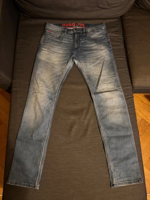 Blå slim fit jeans från Hugo 30/32 - Snygga blå jeans från Hugo i storlek 30/32 med slim passform och klassisk femficksdesign. Jeansen har slitna detaljer och en cool tvättad look, med röd Hugo-logga på linningen och fickan. Perfekta för dig som gillar en smal siluett och streetstyle.