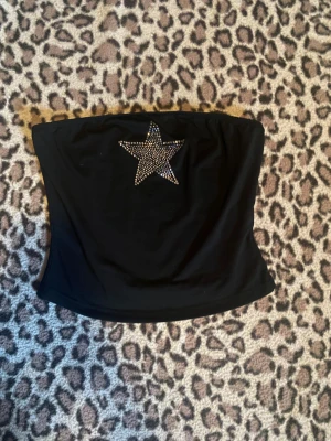 Svart tubtopp med stjärna - Svart bandeau tubtopp med en stor glittrig stjärna framtill i silvriga stenar. Toppen är axelbandslös och har en tight passform. Perfekt för dig som vill sticka ut med en cool detalj. Materialet känns mjukt och stretchigt.