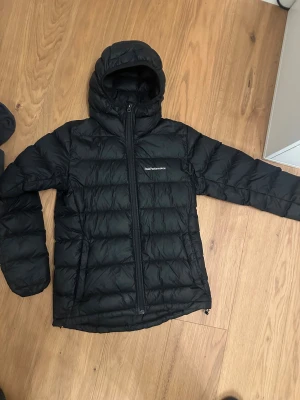 Svart dunjacka Peak Performance S - Svart dunjacka från Peak Performance med huva och quiltad design. Jackan har dragkedja framtill och är perfekt för kalla dagar. Klassisk, stilren look med diskret logga på bröstet. Långärmad och med normal passform.