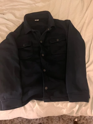 Svart overshirt från Lindex 146-152 - Svart overshirt från Lindex med klassisk krage, två bröstfickor med tryckknappar och raka ärmar. Jackan har en stilren look och är gjord i mjukt bomullsmaterial. Perfekt för lager-på-lager och enkel att matcha med olika outfits.