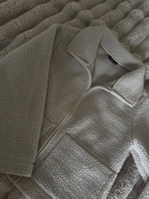 Beige teddyjacka från 157, storlek S - Mysig beige teddyjacka från 157 med krage och hel dragkedja framtill. Jackan har två stora fickor och är tillverkad i fluffigt syntetmaterial som ger en chill och varm vibe. Perfekt för kalla dagar när du vill vara både bekväm och snygg.