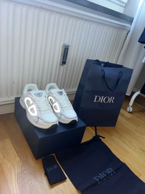 Vita Dior b30 sneakers med reflekterande detaljer - Säljer ett par snygga vita Dior sneakers med reflekterande CD-logga på sidorna. Skorna har rund tå, snörning och platt sula. Materialet är en mix av skinn och syntet, och de kommer med originalförpackning, dustbag och påse från Dior. Perfekt för dig som gillar exklusiva märken och stilrena detaljer.