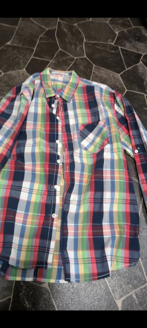 Färgglad rutig skjorta med knappar stl L - Säljer en riktigt färgglad skjorta med rutigt mönster i blått, rött, grönt, gult och vitt. Skjortan har klassisk krage, långärmade ärmar och vita knappar framtill. En bröstficka med samma rutiga mönster ger extra stilpoäng. Perfekt för dig som gillar att sticka ut.