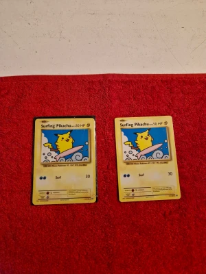 Surfing Pikachu (BUDA!!!) - Två coola Pokémonkort med Surfing Pikachu! Perfekt för dig som älskar Pokémon och vill utöka din samling med en ikonisk och lekfull Pikachu på surfbräda. Samlarkort som passar både nya och erfarna fans.