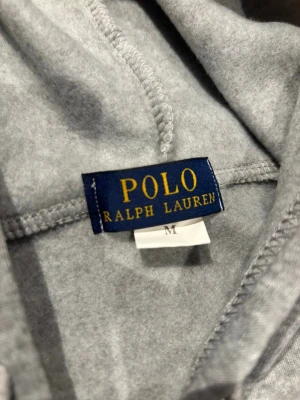 Grå hoodie från Polo Ralph Lauren M - Snygg grå hoodie från Polo Ralph Lauren i storlek M. Klassisk zip-modell, broderad logga på bröstet och kängurufickor. Mjuk bomullsblandning som är skön att ha på sig. Perfekt för en chill och stilren look.