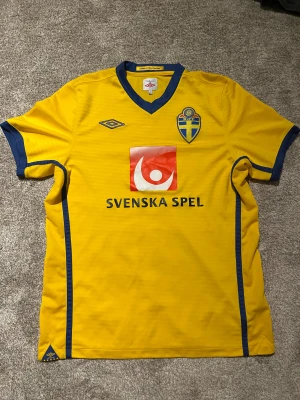 Svenska landslagets gul Umbro fotbollströja från 2010-2011. Riktig  - Gul fotbollströja från Umbro med blå detaljer, officiell Svenska landslaget-design. Tryck med Svenska Spel och Oddset, samt SVFF-emblem på bröstet. V-ringad hals och korta ärmar. Materialet är lätt och andas, perfekt för match eller träning. Använd enstaka gånger