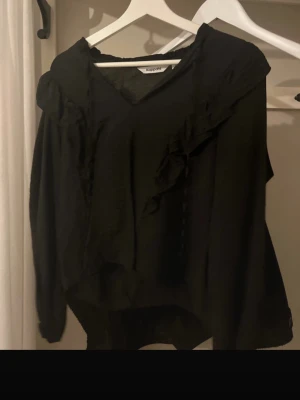 Svart blus med volang från Kappahl - Stilren svart blus från Kappahl med v-ringning och volangdetaljer längs axlarna. Blusen har lång ärm och är tillverkad i ett tunt, lätt material som ger en luftig känsla. Perfekt att matcha med jeans eller kjol för en trendig look.