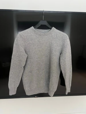 Grå garde Grisch stickad tröja i ullmix - En stilren grå stickad tröja med rund hals och långa ärmar. Tröjan har ribbade muddar vid ärmslut, halsringning och nederkant. Perfekt basplagg i mjuk ullmix som passar till allt. Oanvänd 