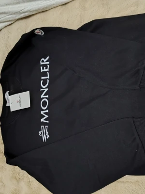 Svart sweatshirt från Moncler - Snygg svart sweatshirt från Moncler med stort vitt Moncler-tryck på bröstet och klassisk Moncler-logga på ärmen. Tröjan har rund halsringning och ribbade muddar vid ärmslut och nederkant. Perfekt för en clean och stilren look.