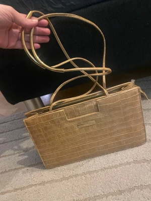 Beige handväska i krokopräglat skinn  - Stilren beige handväska  i krokopräglat skinn. Väskan har rektangulär form, smala handtag och en metallbricka. Köpt sacond hand väldigt vintage är den. Tror ej äkta och små defekter inne som inte syns  därav priset. Använd gärna köp nu❤️