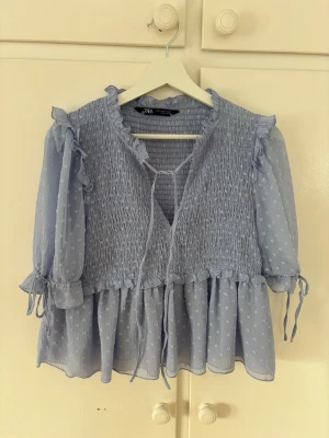 Ljusblå blus med volanger från Zara - Supersöt ljusblå blus från Zara med smockad överdel, volangdetaljer och knytband fram. Blusen kortärmad med små knytband vid ärmsluten och är gjord i ett tunt, prickigt tyg. Storlek S🩷