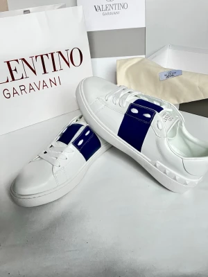 Valentino Garavani vita sneakers - Snygga vita sneakers från Valentino Garavani med en bred blå detalj över mitten och vita nitar på hälen. Skorna har snörning och är tillverkade i slätt skinn för en clean och modern look. Perfekta för dig som vill sticka ut med stilrena detaljer.