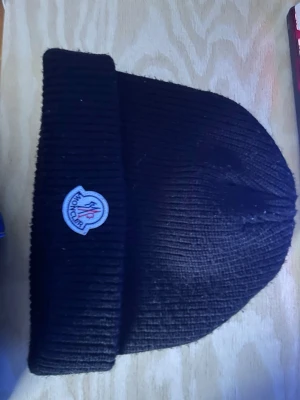 Svart ribbstickad mössa från Moncler - Snygg svart ribbstickad mössa från Moncler med uppvikt kant och klassisk Moncler-logga framtill. Tillverkad i mjukt material som håller värmen och ger en clean look. Perfekt accessoar för kyliga dagar och enkel att matcha med olika outfits.