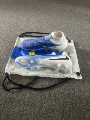 Nike Phantom blåa fotbollsskor - Säljer ett par Nike Phantom fotbollsskor i blått och vitt med gula detaljer. Skorna har en strumpliknande passform och är tillverkade i syntetmaterial för bra grepp och kontroll på planen. Sulan har dobbar för spel på gräs. Snygg och modern design. Använda 2-3 gånger.