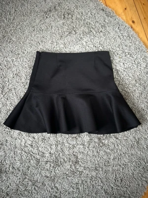 Svart volangkjol med utsvängd nederdel - Snygg svart kortkjol med volang nertill och figurnära passform. Säljer då den är för stor för mig! Skickar snabbt