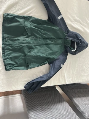 Grön regnjacka med huva - Snygg grön regnjacka med mörkblå ärmar och huva. Jackan har tryckknappar framtill, två stora fickor med lock och reflexdetaljer på ärmarna. Perfekt för regniga dagar och enkel att matcha med din outfit.