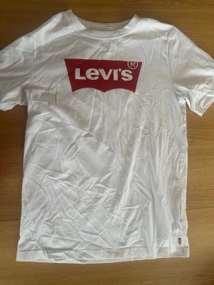 Vit Levi's t-shirt med röd logga - Klassisk vit t-shirt från Levi's med den ikoniska röda loggan tryckt på bröstet. T-shirten har rund halsringning och korta ärmar. Tillverkad i mjuk bomull som är skön att bära. Perfekt till jeans eller shorts för en enkel och snygg look. Passar ungefär s.