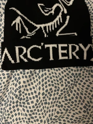 Svart mössa från Arc'teryx - Snygg svart mössa från Arc'teryx med vit logga och text framtill. Klassisk uppvikt kant och stickad design som ger en clean och sportig look. Perfekt för kalla dagar och outdoor vibes.