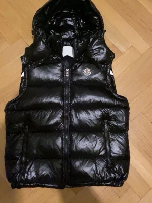 Svart dunväst från Moncler med huva - Snygg svart dunväst med glansig finish och avtagbar huva. Aldrig använd!!! Västen har två dragkedjeförsedda fickor framtill och Moncler-logga på bröstet. Perfekt för lager-på-lager och streetwear-vibe.
