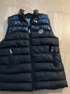Svart dunväst från Moncler - Snygg svart dunväst från Moncler med glansig finish upptill och klassisk logotyp på bröstet. Västen har två dragkedjeförsedda fickor och hög krage. Perfekt för lager-på-lager och streetstyle. Materialet är quiltat och fyllt med dun för extra värme.