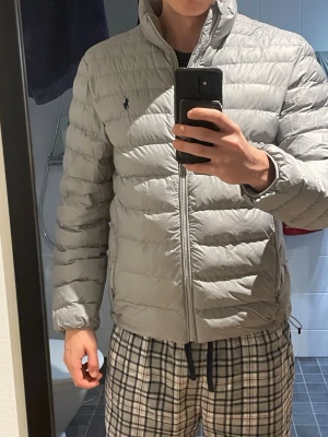 Ljusgrå dunjacka från Polo Ralph Lauren - Snygg ljusgrå dunjacka från Polo Ralph Lauren med klassisk pufferdesign och diskret logga på bröstet. Jackan har dragkedja framtill, hög krage och två sidofickor med dragkedja. Perfekt för kyliga dagar och enkel att matcha.