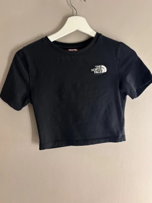 Svart croppad topp från The North Face - Snygg svart croppad topp från The North Face med korta ärmar och rund hals. Diskret logga broderad på bröstet. Tillverkad i mjuk bomull som känns skön mot huden. Perfekt till jeans eller kjol för en avslappnad look.
